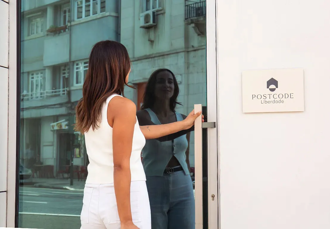 Mujer de espaldas abre una puerta de cristal reflejante; cartel dice "POSTCODE Liberdade".