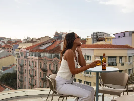 Mujer sentada en una terraza con bebida, mirando la ciudad al atardecer.