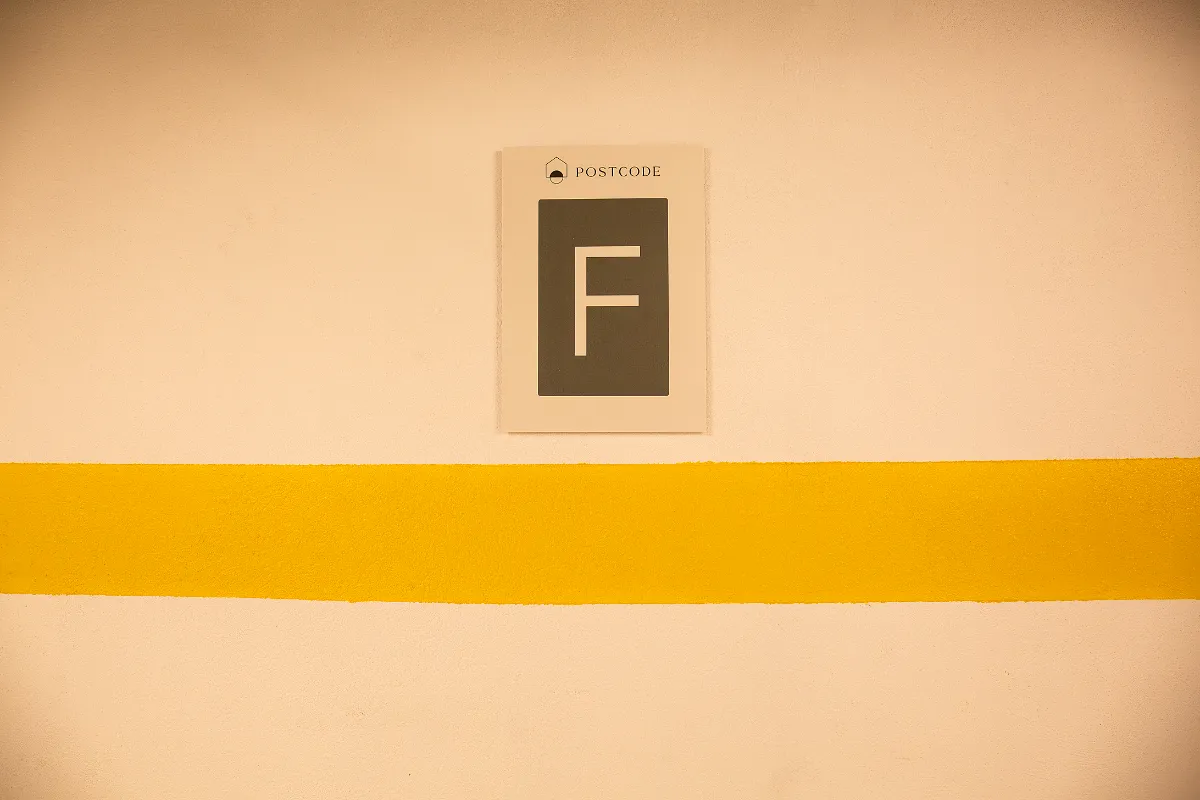 Señal gris con letra "F" y la palabra "POSTCODE" sobre fondo amarillo y pared beige.