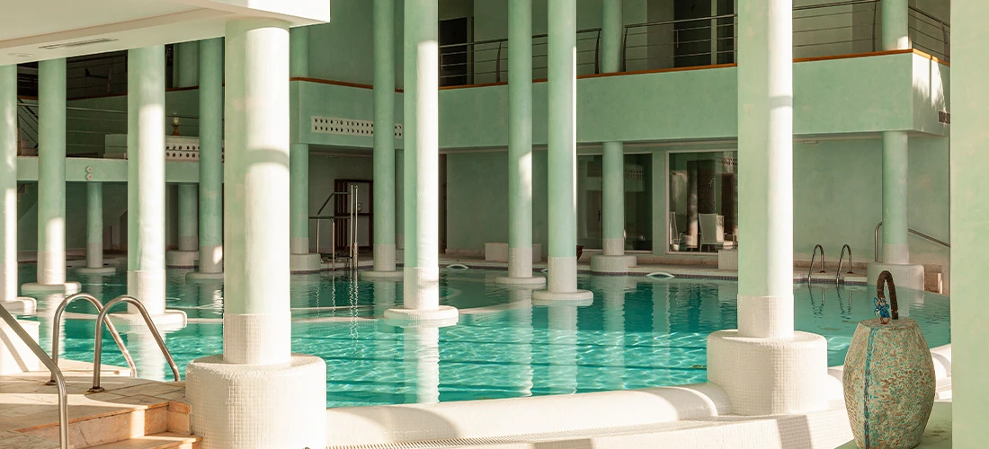Vista lateral de piscina climatizada con escaleras de acceso y columnas verdes.