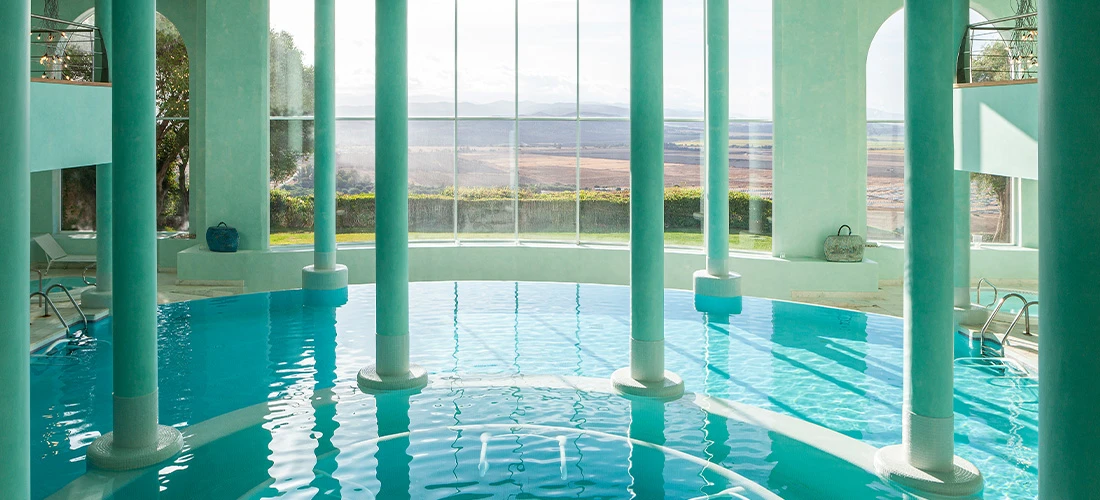 Vista lateral de piscina climatizada con escaleras de acceso y columnas verdes.
