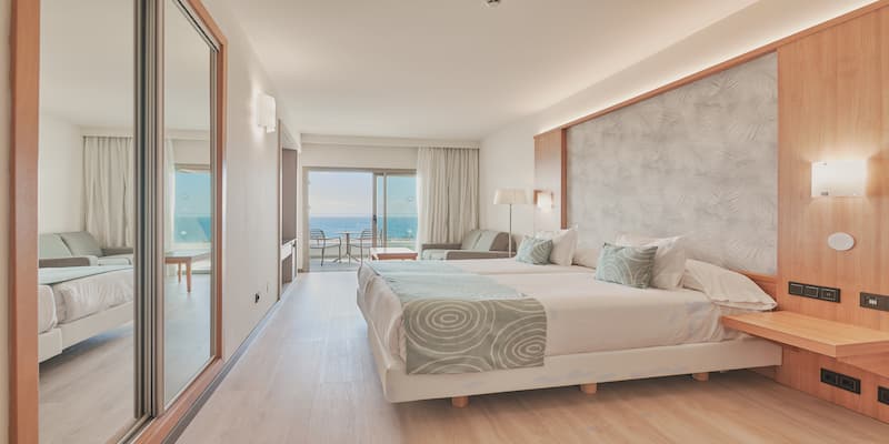Habitaci&oacute;n moderna de hotel con cama grande, sof&aacute; y balc&oacute;n con vista al mar.