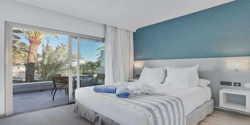 Habitaci&oacute;n de hotel moderna con cama, balc&oacute;n y vista a palmeras.