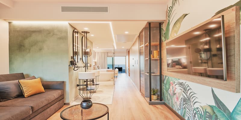Sala moderna con sof&aacute; marr&oacute;n, coj&iacute;n amarillo, mesa de cristal y decoraci&oacute;n tropical en la pared.