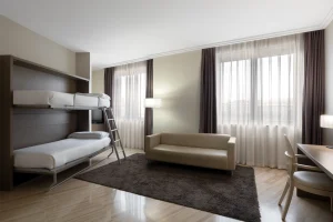 A modern hotel room featuring a bunk bed setup, cozy sofa, and large windows with sheer curtains.