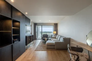 A modern living room featuring a gray sectional sofa, wooden flooring, and large windows with a view.
