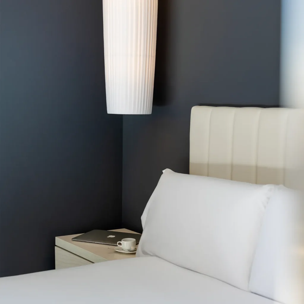 A laptop and coffee cup sit on a nightstand next to a neatly made bed