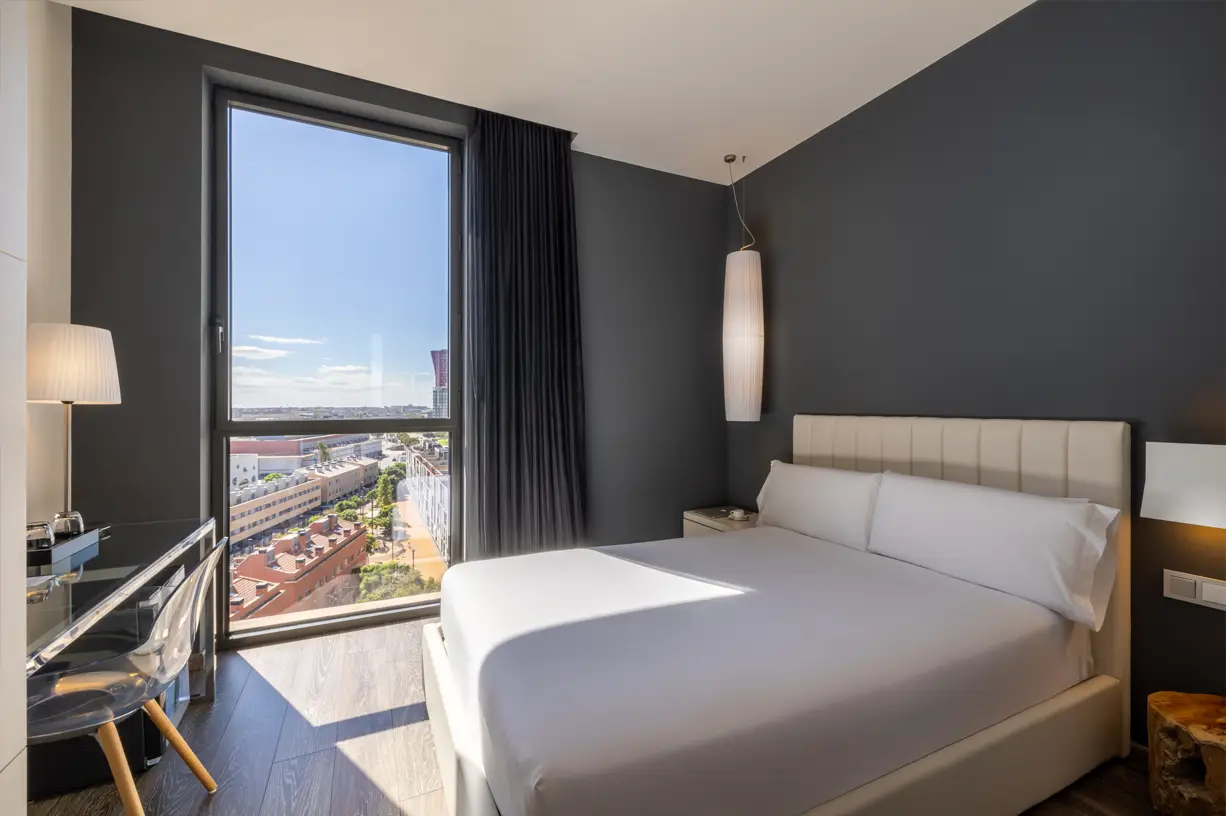 A room with a large window offering a city view and a comfortable bed