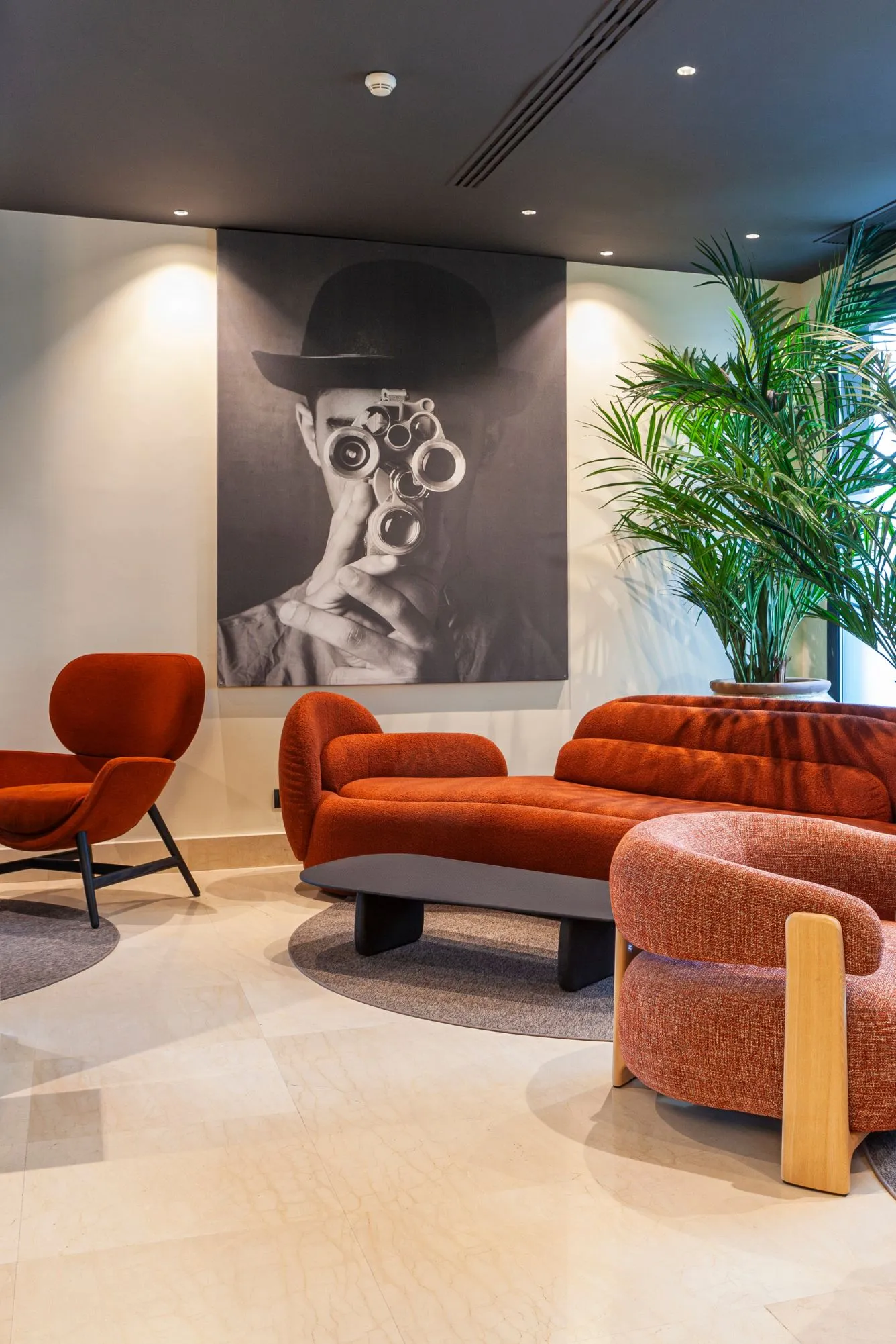 A living area with orange sofas, a large black and white portrait of a man, dim lighting, and a tall indoor plant