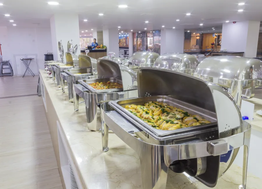 Mesa de buffet con recipientes de comida y aparadores inox en salón iluminado.