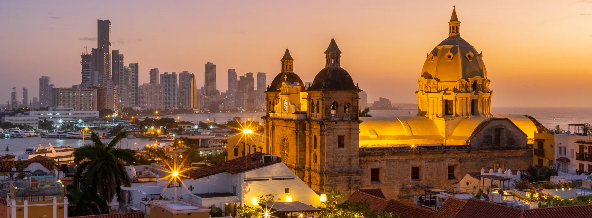 Catedral y ciudad costera al atardecer, luces nocturnas y rascacielos al fondo