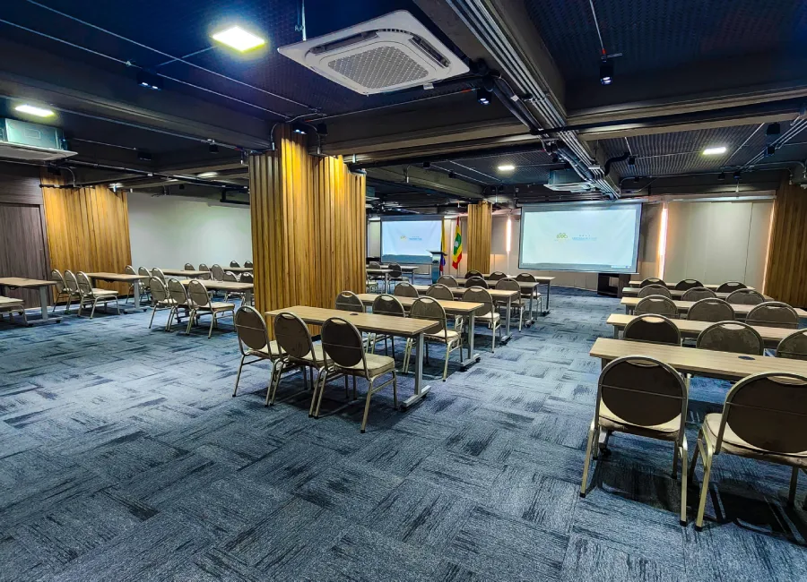 Sala de conferencias con mesas, sillas y pantalla de proyecci&oacute;n al fondo