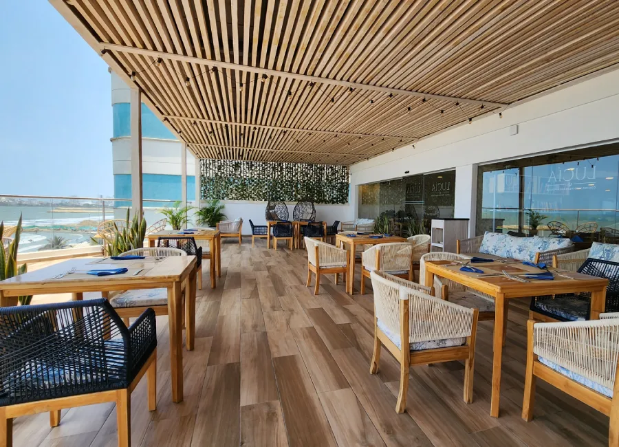 Terraza frente al mar con mesas y sillas bajo techo de madera y cortinas de plantas
