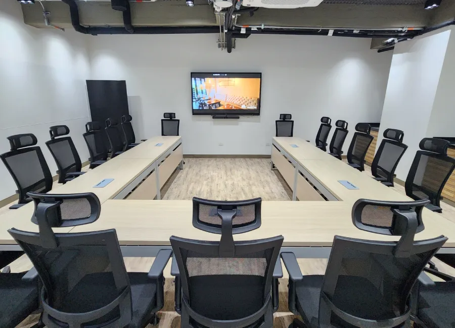 Sala de reuniones moderna con mesas y sillas, pantalla al fondo y proyector.