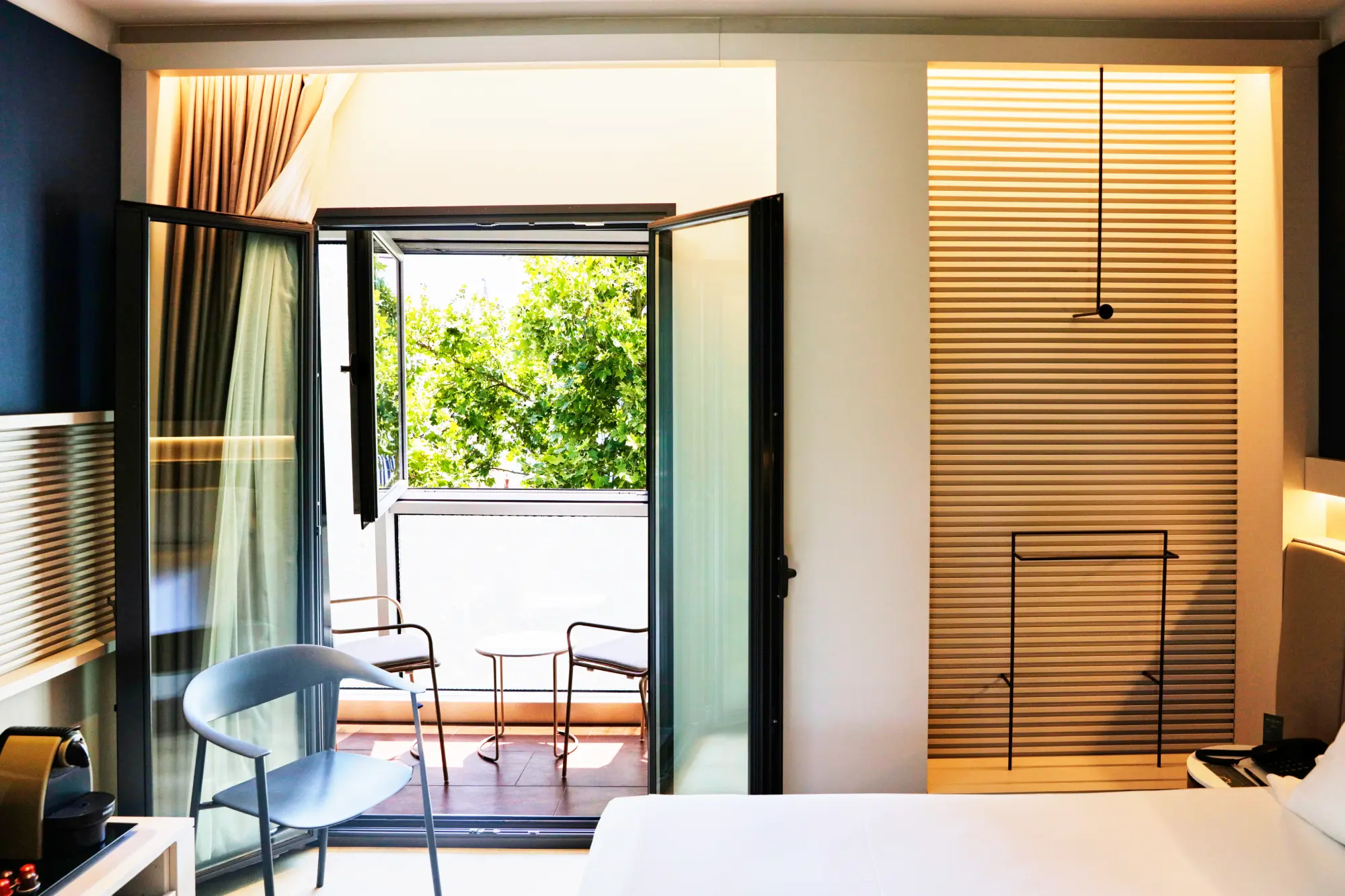Habitación moderna con balcón, sillas y mesa, vista a árboles verdes, y decoración minimalista.