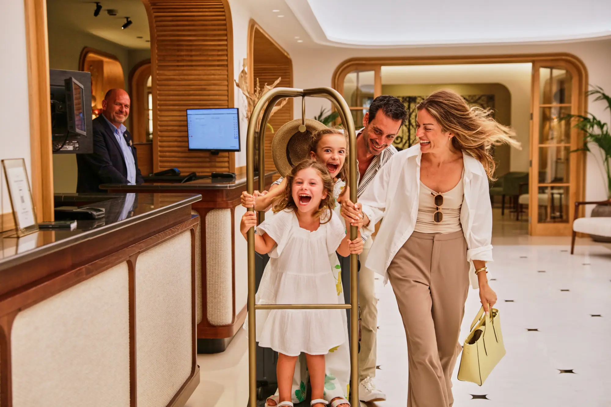 Familia feliz entra a un hotel con niños jugando en el carrito de equipaje, recepcionista sonriente.