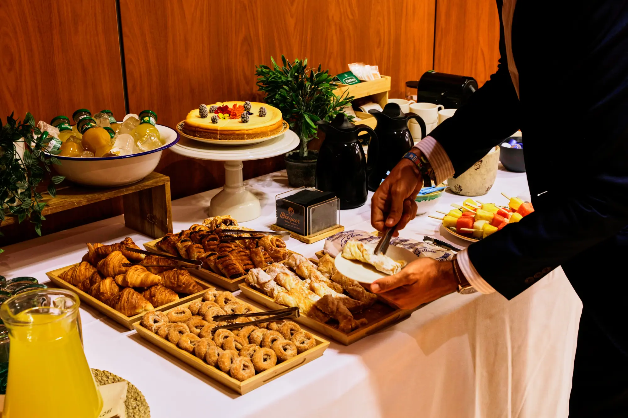 Mesa de buffet con repostería, frutas, jugos y café; una persona sirve un pastel en su plato.