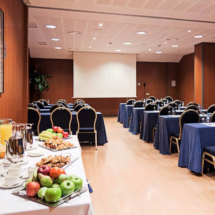 Sala de conferencias con mesas, sillas y mesa de refrigerios con frutas y jugo.