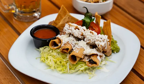 Plate of rolled tacos with toppings and a side of sauce on a wooden table.