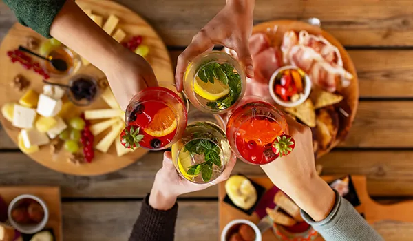 Hands toasting with colorful drinks over a table of appetizers.