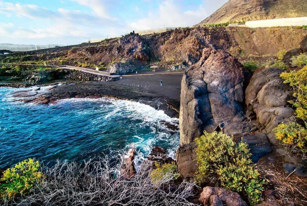Costa rocosa con playa de arena negra y olas rompiendo, rodeada de vegetación y acantilados.