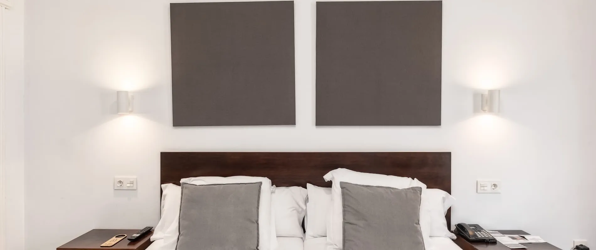 Cama con dos cojines grises, adornada con cuadros gris oscuro en la pared, lámparas a los lados.