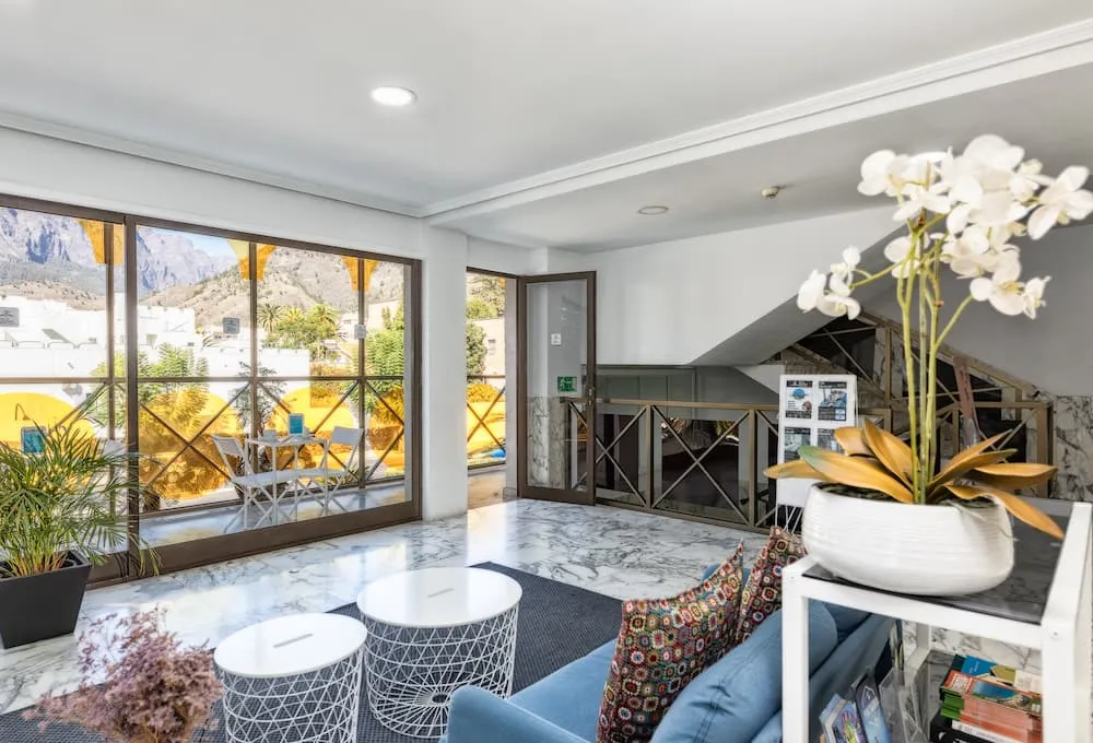 Sala con mesa, sillón azul y planta; fondo con ventanas grandes y vista a las montañas.