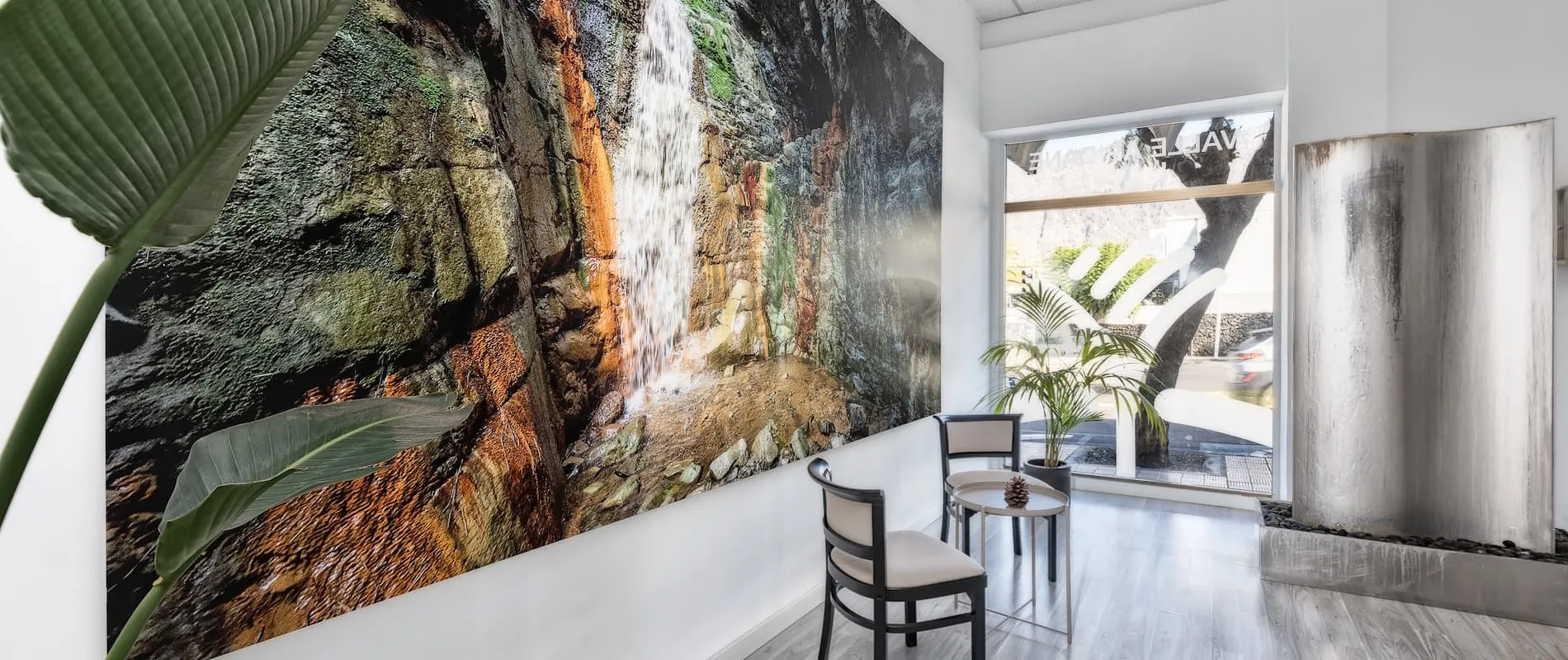 Sala de espera con cuadro de cascada, dos sillas, mesa y plantas cerca de una ventana.