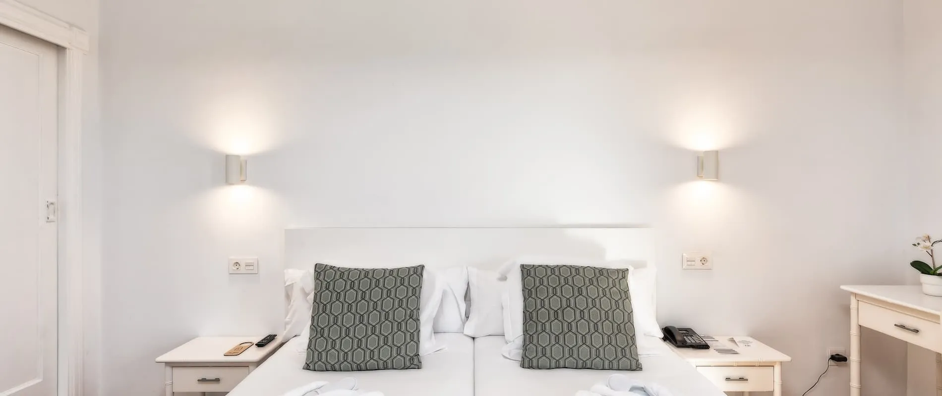 Cama blanca con cojines grises, mesas de noche con lámparas, teléfono y planta decorativa.