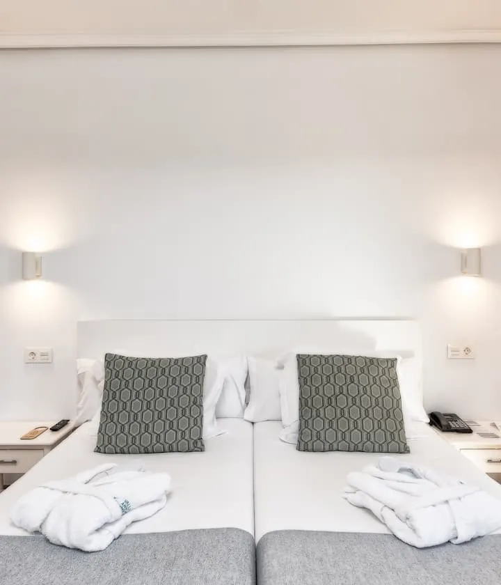 Dos camas blancas con cojines grises, batas y lámparas de pared encendidas.