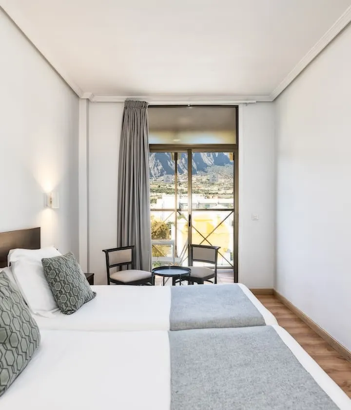 Habitación luminosa con dos camas, ventanas grandes y vista a las montañas.