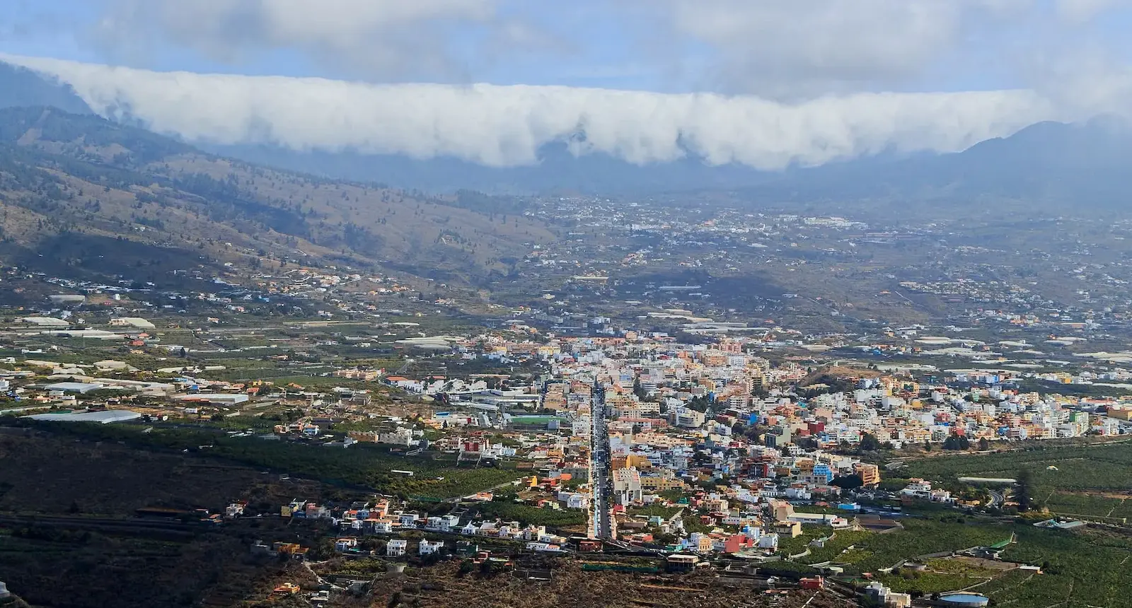 Vista panorámica de un valle con un pueblo, rodeado de montañas y nubes bajas en el fondo.