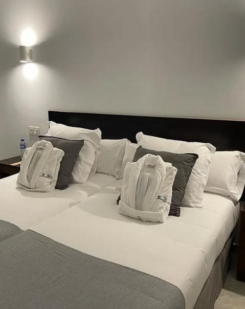 Habitación con cama doble, cojines grises y albornoces sobre la cama, con luz suave de pared.