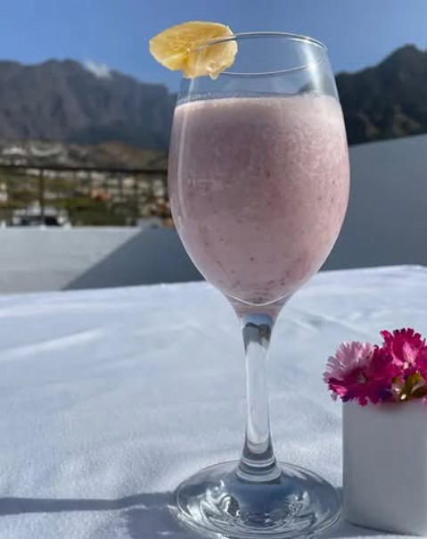 Copa de batido rosado con piña en el borde, flores rosa al lado, montaña de fondo.
