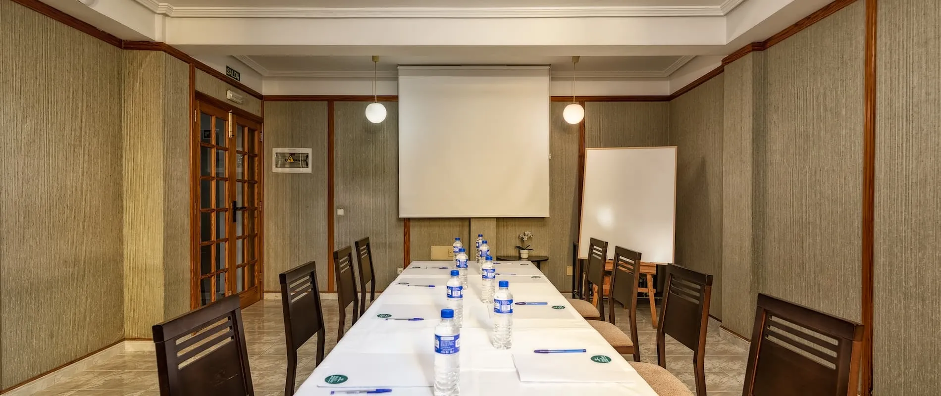 Sala de conferencias con mesa rectangular, sillas, proyector y pizarra blanca.