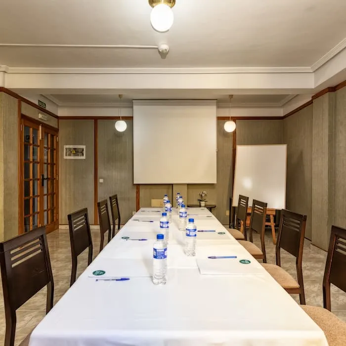 Sala de reuniones con mesa larga, sillas, botellas de agua, bolígrafos y proyector.