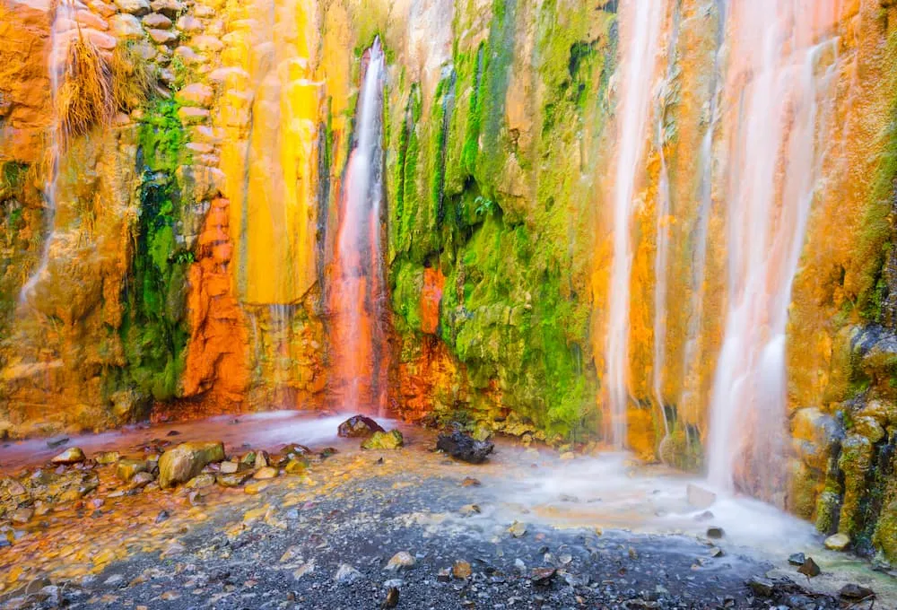 Cascada de colores vivos en un muro de piedra con musgo y rocas al pie, agua fluye suavemente.