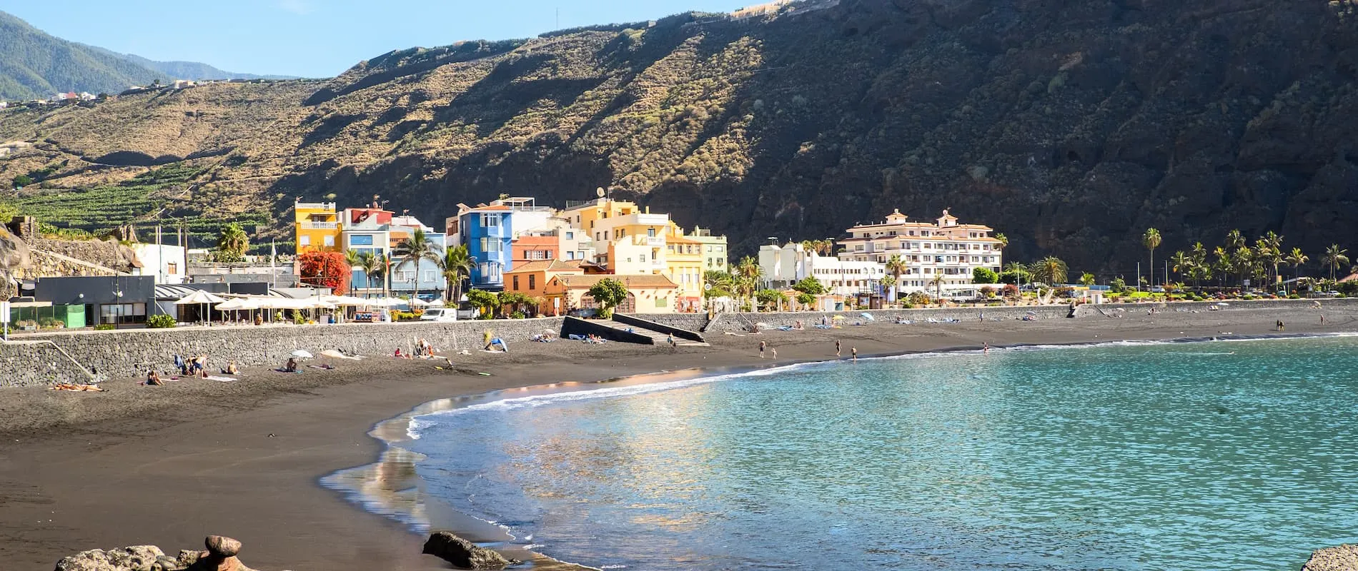 Playa de arena negra con casas coloridas y montañas al fondo.