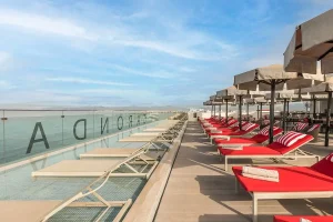 Red sun loungers and umbrellas on a rooftop deck with a pool and blue sky.