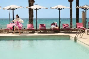 Poolside scene with a woman twirling and a man relaxing on pink loungers by the sea.