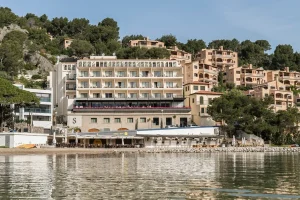 Building by the water with hillside houses surrounded by trees in the background.