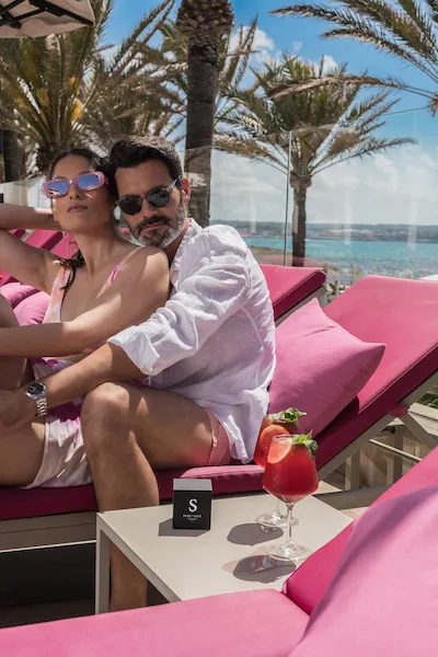 Couple relaxing on pink loungers by the beach with cocktails under palm trees.