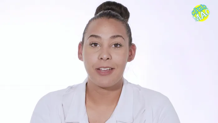 Person in a white top with "Club MAC" logo; neutral expression, bun hairstyle.