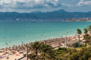 Sunny beach with colorful umbrellas, people swimming, palm trees, and distant mountains under a cloudy sky.