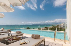 Sun loungers by a pool with a sea view, under umbrellas, with drinks and an open book nearby.