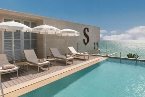 Rooftop pool with lounge chairs and umbrellas overlooking the ocean, with a large "S" on the building wall.