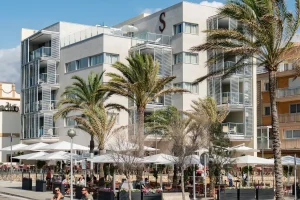 Modern building with balconies, palm trees, and outdoor seating under umbrellas on a sunny day.