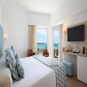 Hotel room with sea view, double bed, TV, and two chairs by the window.