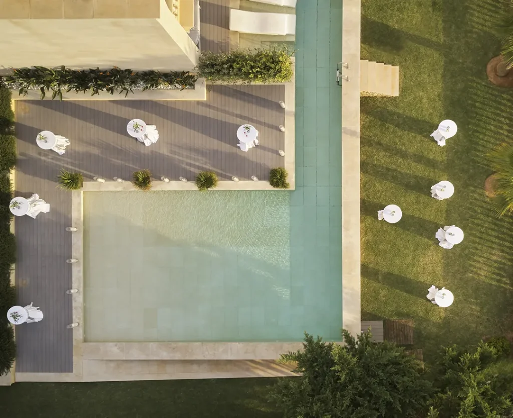 Aerial view of a pool and garden with tables and chairs arranged on grass and deck.