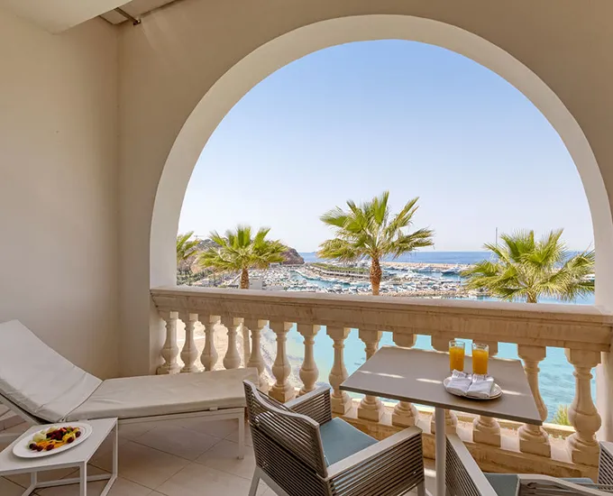 Balcony with lounge chairs, table and ocean view through an archway with palm trees.